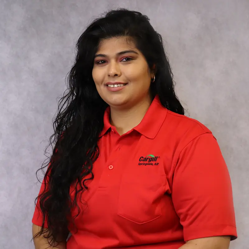 A woman with long black hair smiles while wearing a red collared shirt with a logo on it. The background is a light gray, creating a professional portrait setting.