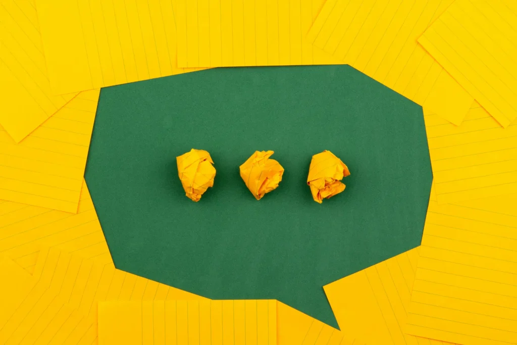 Three crumpled yellow paper balls placed inside a green speech bubble shape, set against a bright yellow background with lined paper sheets.
