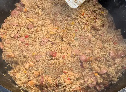 A massive black cast-iron pot filled with a hearty Cajun jambalaya containing seasoned rice, sliced smoked sausage, and pieces of chicken being stirred with a large metal spatula.