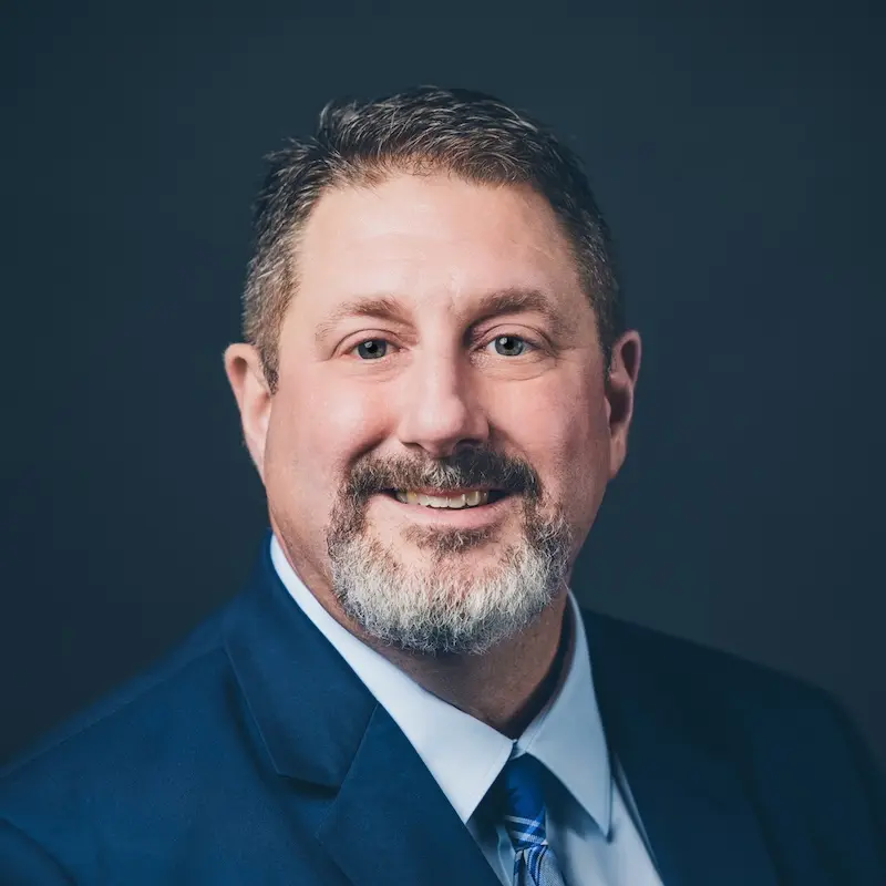 Shane Acosta: Headshot of Shane Acosta, Founder of Down The Bayou Meats, wearing a blue suit and tie against a dark background.