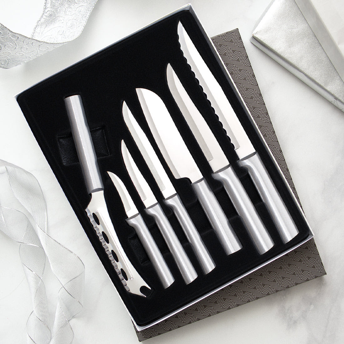 A black gift box containing a six-piece set of silver-handled kitchen knives. The set includes a cheese knife with cutouts, two small paring knives, a utility knife, a slicing knife, and a long serrated bread knife, displayed next to a silver ribbon on a marble surface.