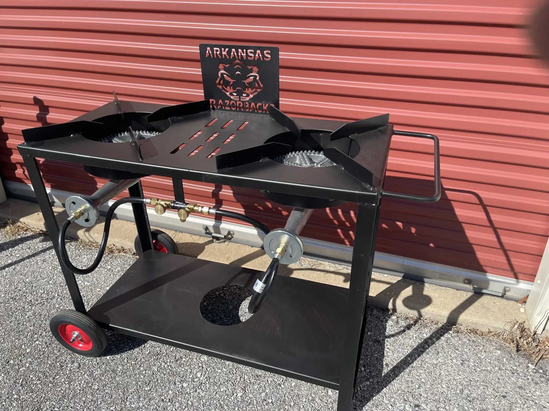 A black metal outdoor double-burner gas cart. It features a custom backplate with the Arkansas Razorbacks logo and team name, a lower shelf with a circular cutout for a propane tank, a side handle, and two wheels with red hubs.