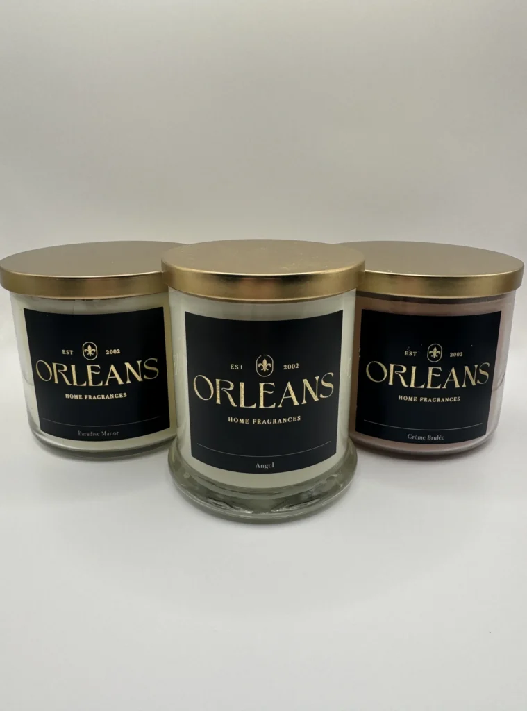 Three Orleans Home Fragrances candles in glass jars with gold lids. The labels show the scents Paradise Manor, Angel, and Crème Brûlée.