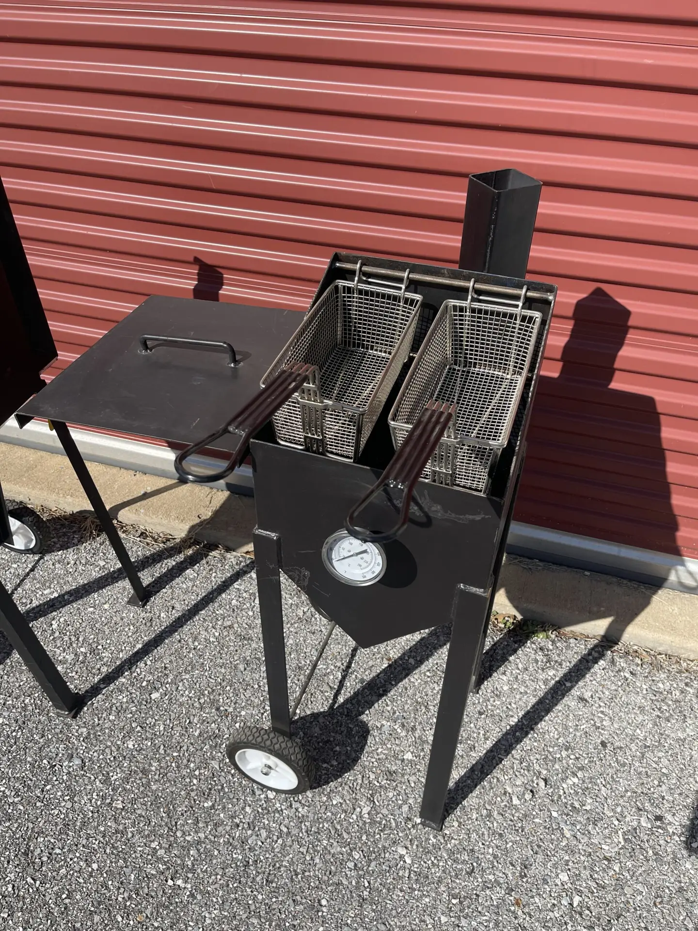 A black metal outdoor deep fryer setup with two rectangular wire frying baskets resting inside. It includes a built-in temperature gauge on the front, a side prep shelf, and wheels for mobility.
