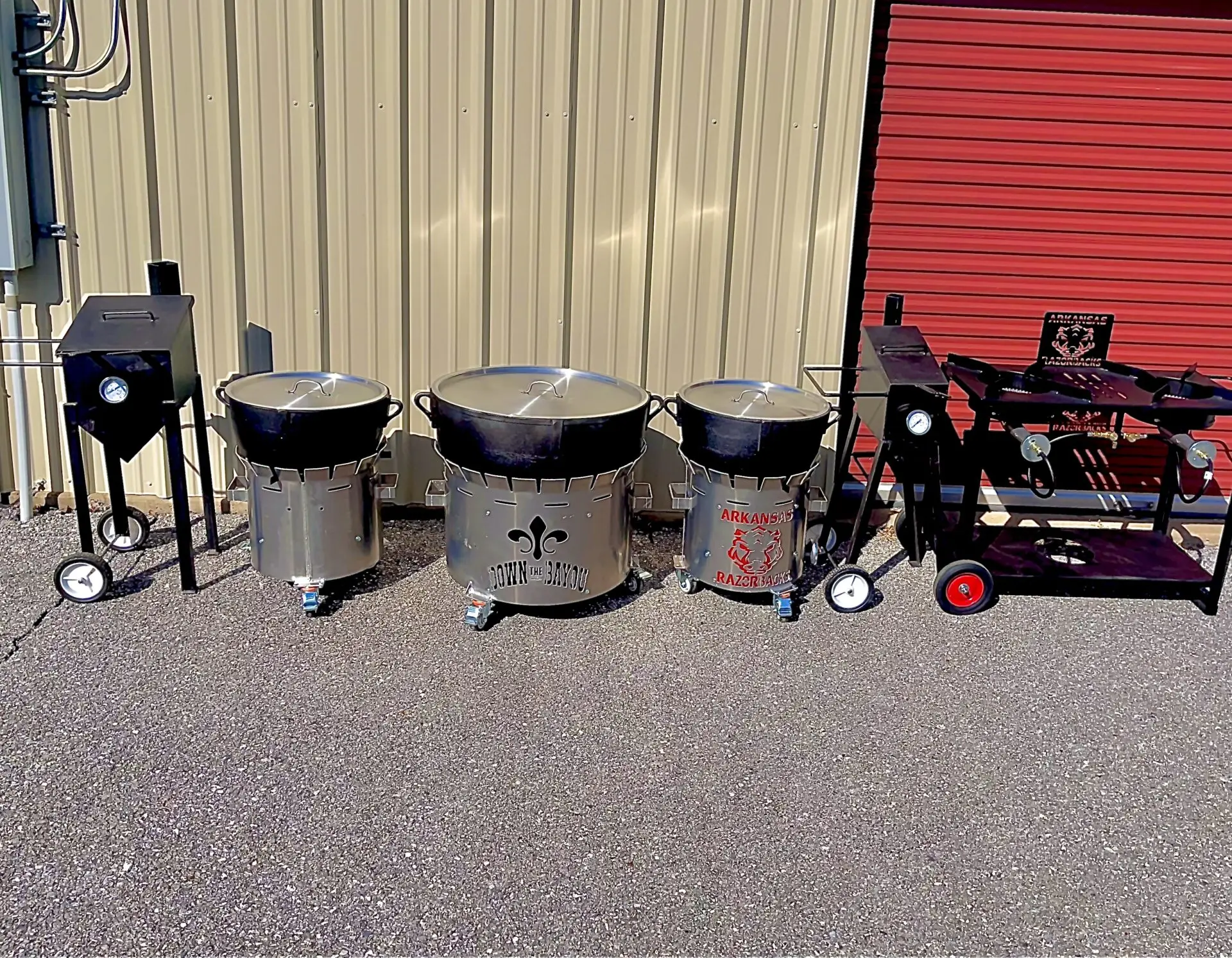 A wide shot showing a lineup of outdoor cooking equipment. From left to right: a single deep fryer, three large cast-iron pots on custom stainless steel burner stands, and a large flat-top outdoor gas stove setup.