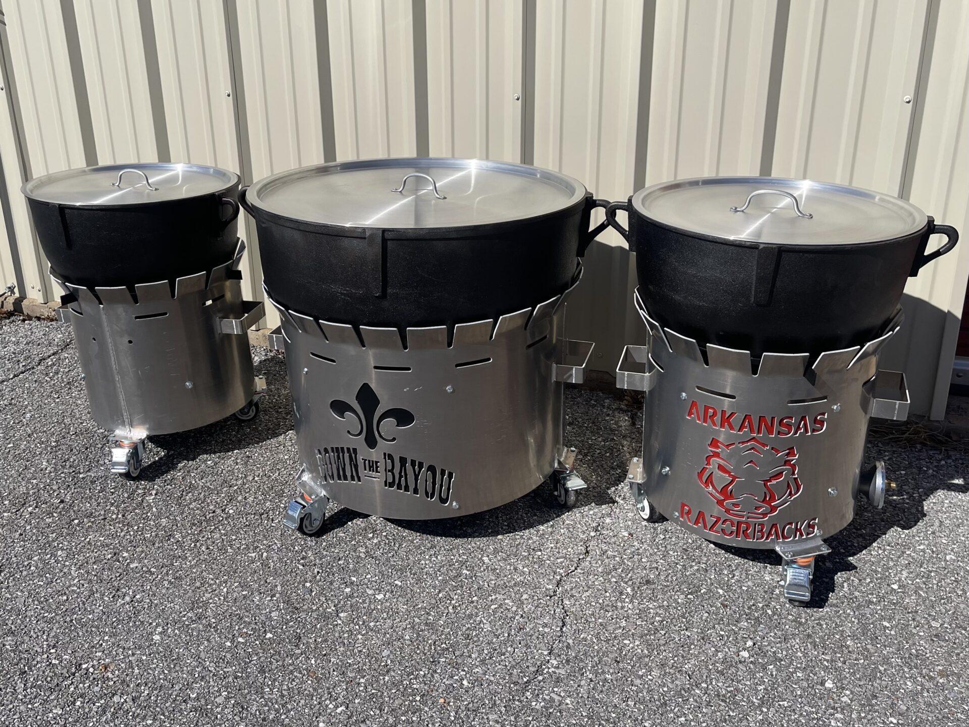 Three large cast-iron cooking pots resting on custom stainless steel burner stands. The middle stand has a Fleur de Lis with the words Down The Bayou, and the right stand features the Arkansas Razorbacks logo.