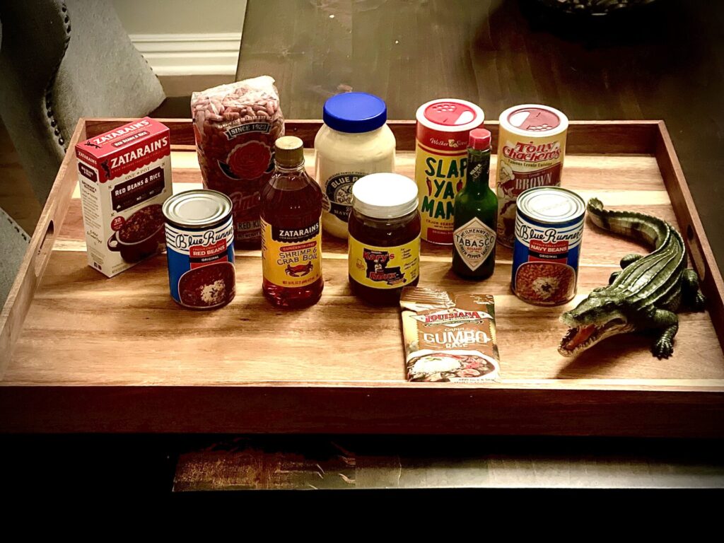 A wooden tray filled with classic Louisiana cooking staples, including Zatarain's fish fry and crab boil, Blue Runner canned beans, Blue Plate mayonnaise, Slap Ya Mama seasoning, Tony Chachere's, and Tabasco, sitting next to a small toy alligator.